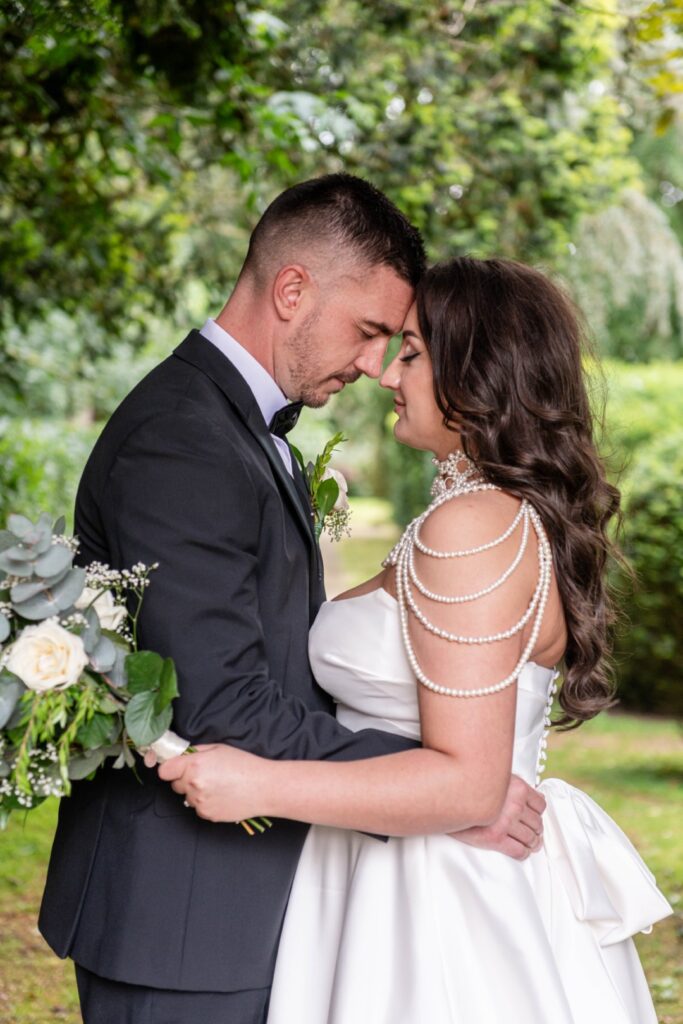 A couple shares an intimate moment, foreheads touching, in the lush green Ansty Hall, with the bride in a pearl-adorned gown and the groom in a classic black tuxedo.