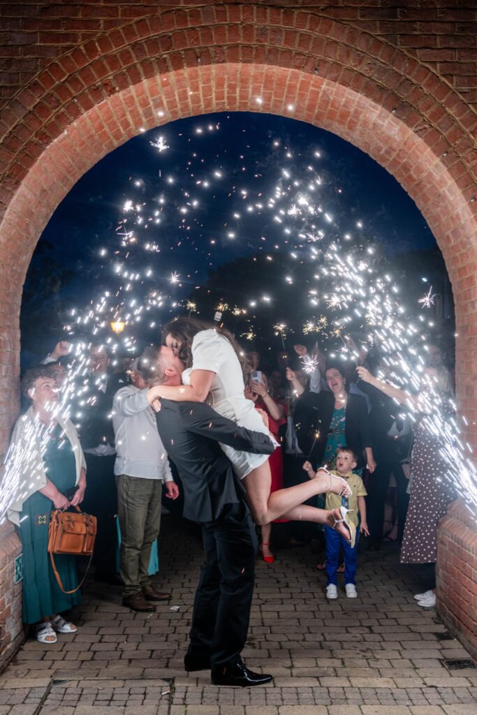 A joyous couple shares a kiss under a shower of sparkler lights, surrounded by cheering guests in a festive atmosphere. Ansty Hall