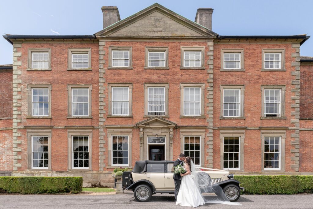 A couple shares a kiss in front of a grand red brick mansion while standing next to a vintage car, capturing a romantic wedding moment - Ansty Hall