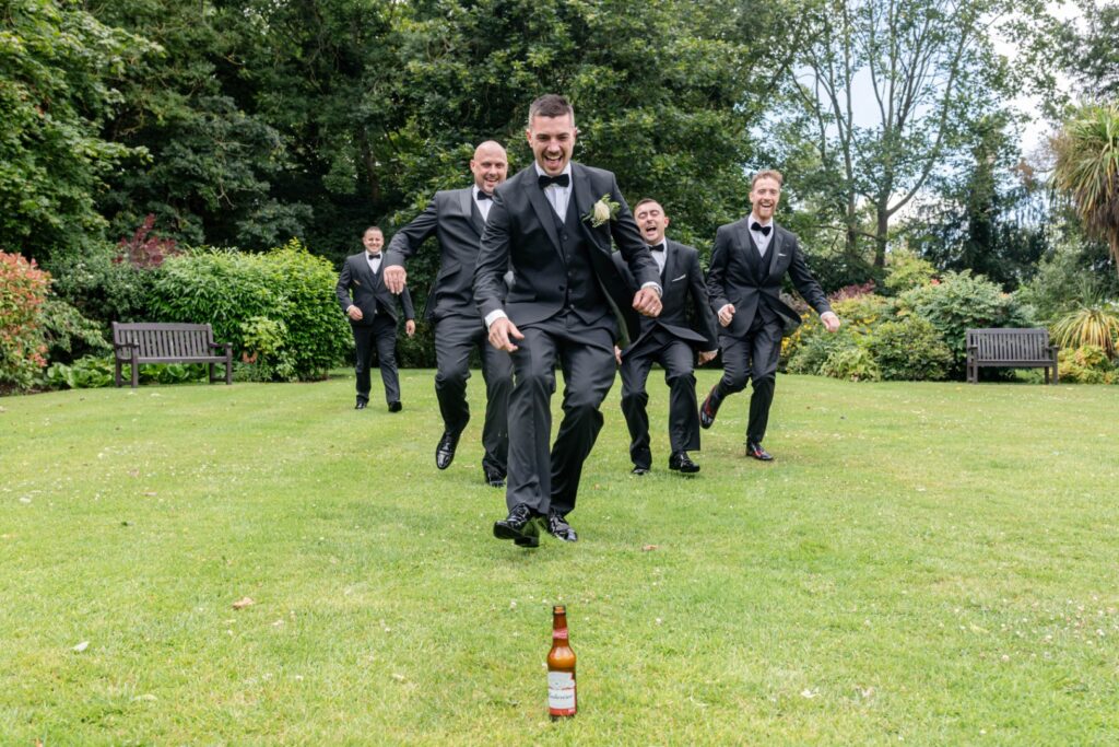 A group of groomsmen in tuxedos joyfully running toward a beer bottle on a grassy lawn, set against a lush garden backdrop. Ansty Hall