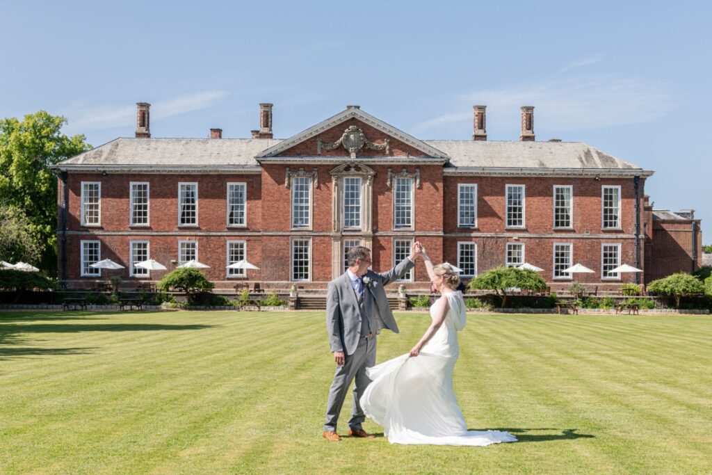 A bride and groom dance playfully on the lawn in front of a beautiful brick building on a sunny day.