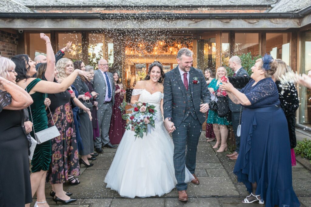 A joyful couple walks hand-in-hand through a crowd of guests showering them with confetti, celebrating their wedding day outside a venue.