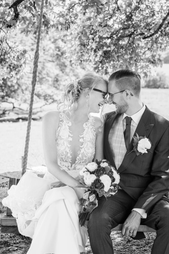 A loving couple sharing a joyful moment on a swing, with the bride holding a bouquet and wearing elegant glasses, while the groom smiles warmly, both surrounded by natural greenery at Ashton Lodge