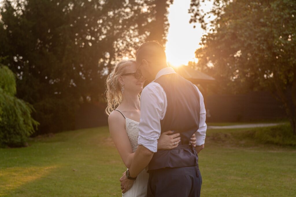 A couple shares a tender moment during sunset, surrounded by lush greenery and warm sunlight.