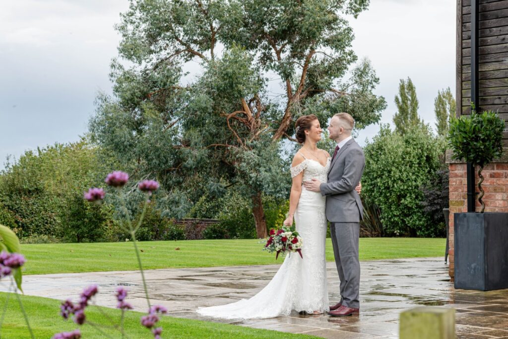 A newlywed couple embracing joyfully outdoors, surrounded by lush greenery and flowers, capturing a heartfelt wedding moment.