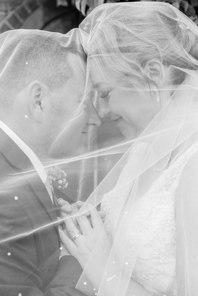 A close-up of a couple sharing an intimate moment while holding hands beneath a wedding veil, conveying love and connection.