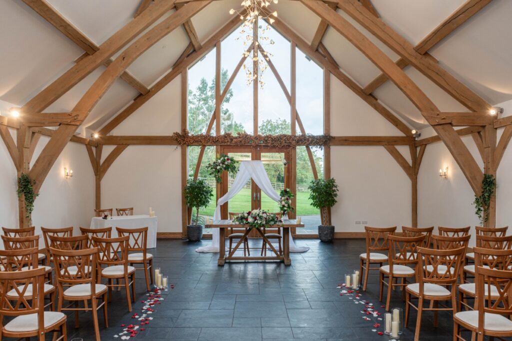 A beautifully arranged wedding setup featuring wooden beams, decorative floral elements, and candles in the bright, spacious Mythe Barn with views of greenery outside.