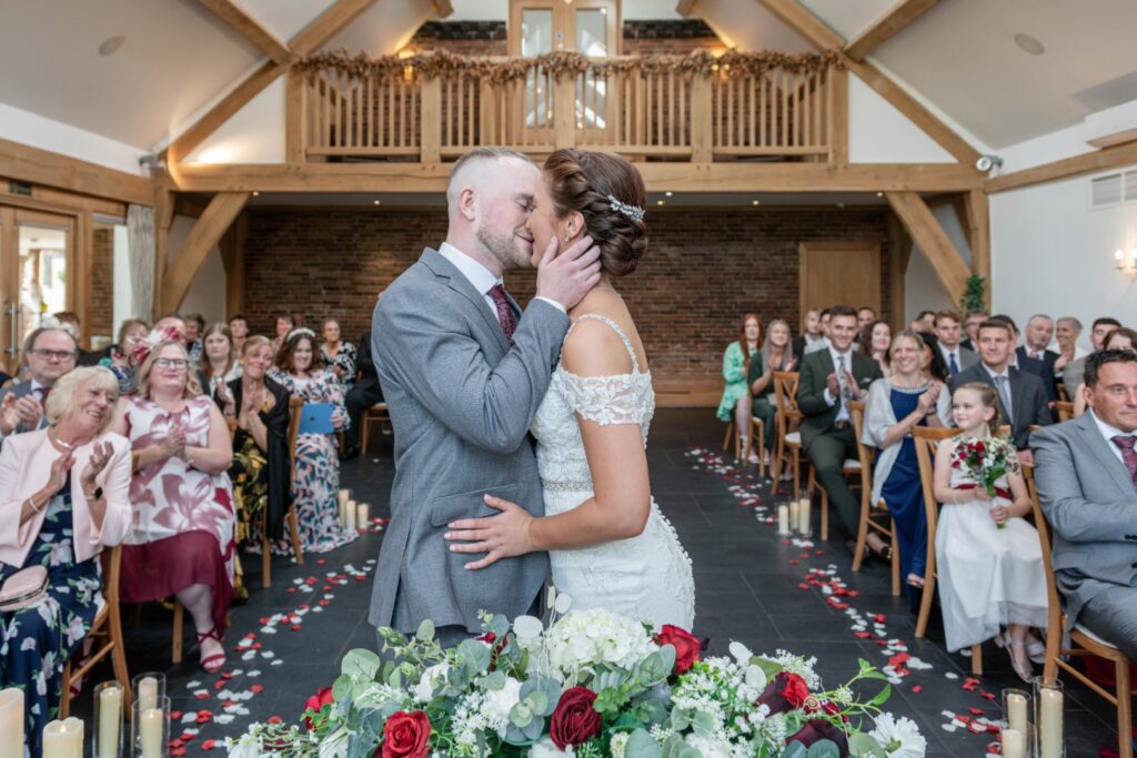 A couple shares a tender kiss during their wedding ceremony, surrounded by joyful guests and floral decorations.