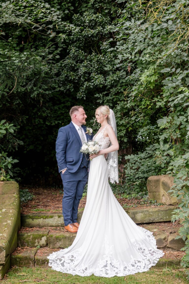 A bride and groom share a joyful moment amidst greenery, with the bride wearing a flowing gown and holding a bouquet of white roses.