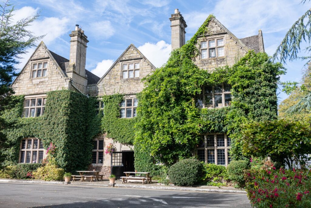 A picturesque view Weston Hall wedding venue near Nuneaton, covered in ivy, featuring multiple gabled roofs and large windows, surrounded by lush greenery and a serene outdoor seating area.