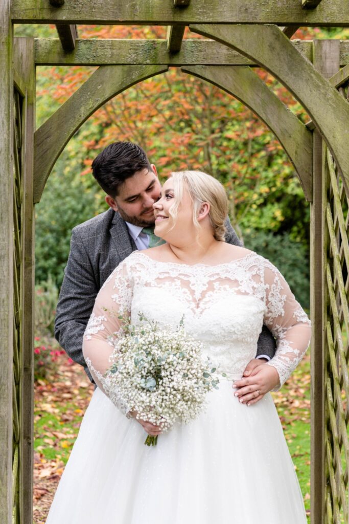 A couple embraces under a wooden arch in a garden, the bride in a lace gown holding a bouquet of flowers, with colorful foliage in the background.