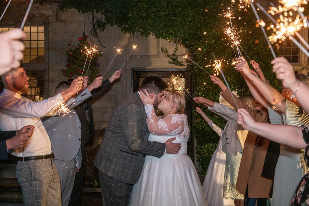 A couple shares a romantic kiss surrounded by guests holding sparklers, celebrating their wedding night in front of Weston Hall