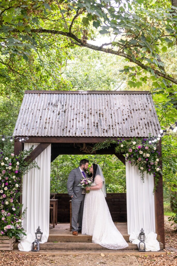 A couple shares a kiss under a rustic gazebo adorned with flowers, surrounded by lush greenery, on their wedding day.