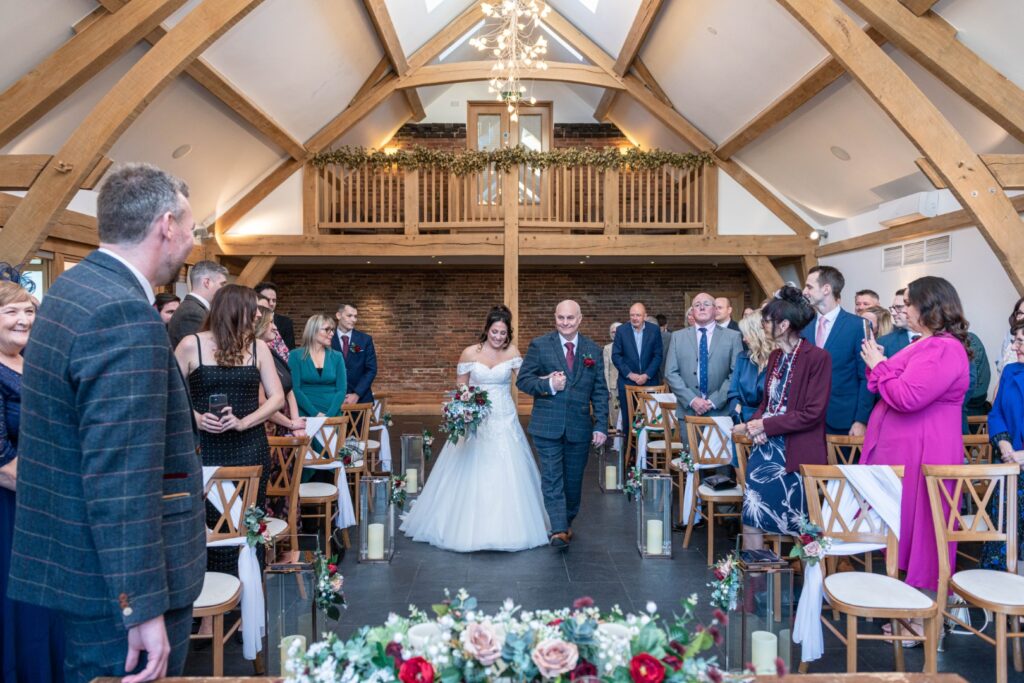 A bride walks down the aisle with her father, surrounded by guests in a beautifully decorated Mythe Barn featuring wooden beams and romantic floral arrangements.