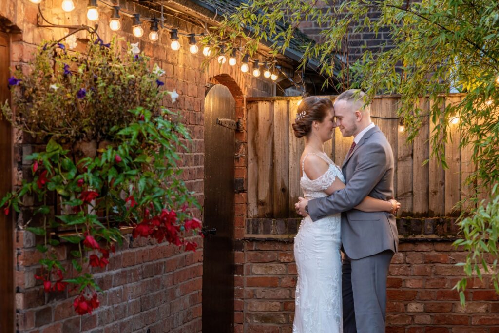 A couple in elegant attire sharing a tender moment in an outdoor setting adorned with string lights and flowers at Mythe Barn