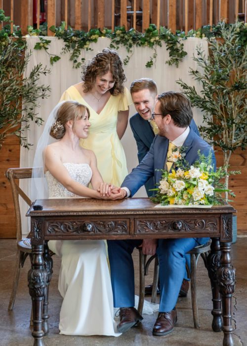 A bride seated holding hands with a groom at a wooden table, while two smiling guests lean in towards them in a rustic setting decorated with green foliage.