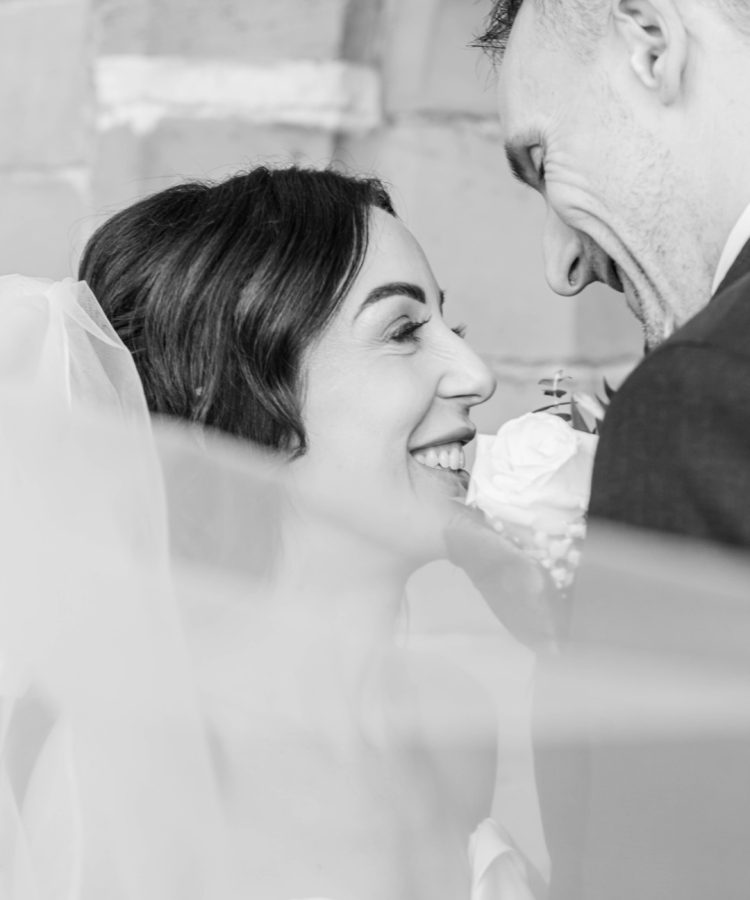 A close-up of a couple smiling at each other, framed by a veil, capturing an intimate wedding moment.