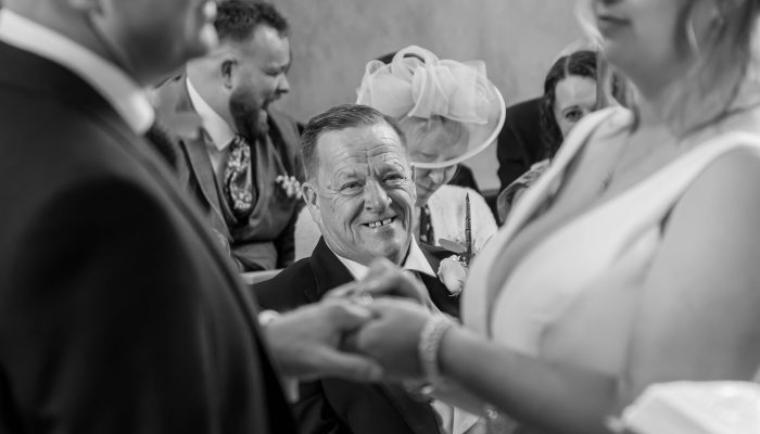 A black and white photo capturing a joyful man smiling as he embraces someone at a wedding ceremony, surrounded by guests.